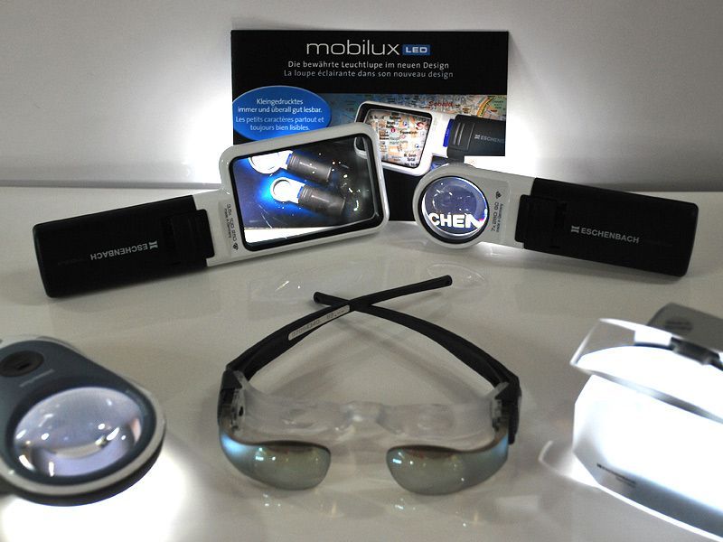 Beleuchtete Lupen und Spezialbrillen auf einem weißen Tisch, im Hintergrund ein Aufsteller mit der Aufschrift mobilux LED von Eschenbach.